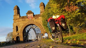 Ein Fahrrad mit rot-weißem Reisegepäck steht vor der Alten Harburger Elbbrücke, das von der Abendsonne in warmes Licht getaucht ist